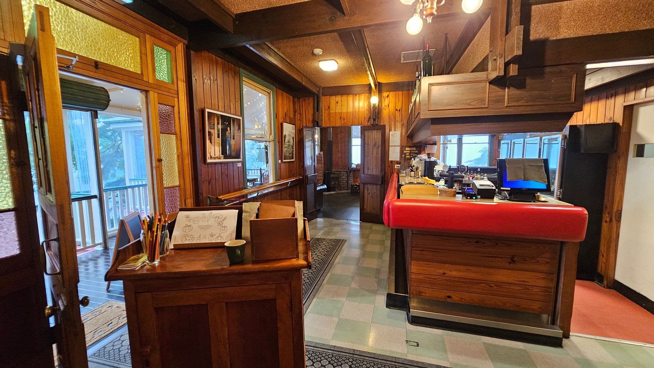 A cosy interior with wooden panelling throughout. On the left, a door with colourful stained glass panels opens to a porch. A small wooden desk holds colouring sheets and pencils. In the centre, a hallway leads to another room, with framed pictures on the walls. On the right, a counter with a red top features a computer screen and various items. The ceiling has exposed beams and hanging lights, while the floor is tiled with a patterned rug near the entrance.