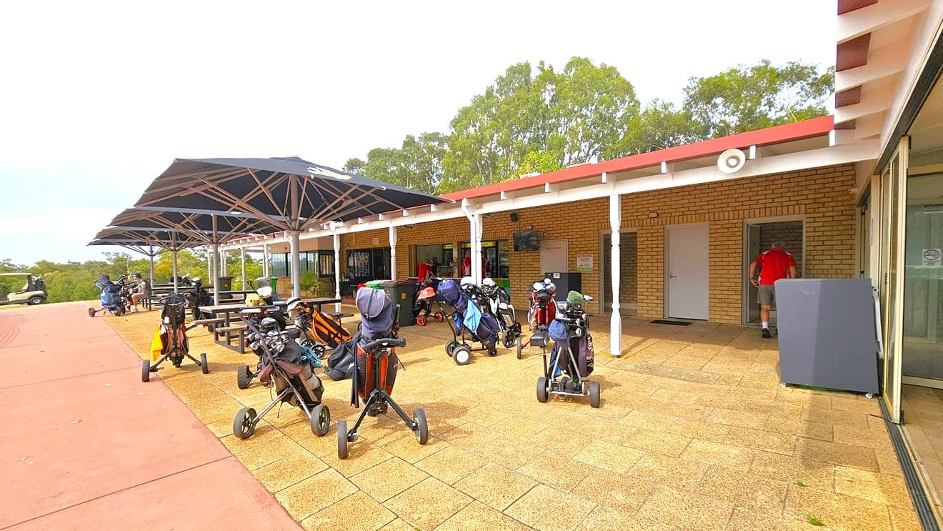 On the right is a brick building housing the cafe. Picnic tables are under a large umbrella shade. Golf trolleys occupy the brick area outside the cafe. To the right is a glass door that leads to the seating area. Far left, a golf cart is visible.