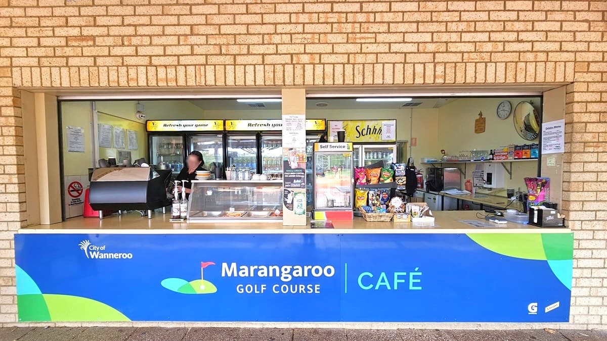 The cafe features a blue banner bearing the facility's name. A food rack atop the counter along with other light snacks. Behind the reception is a fridge for cold drinks. A staff member stands behind the counter. The wall is made of brown brick.