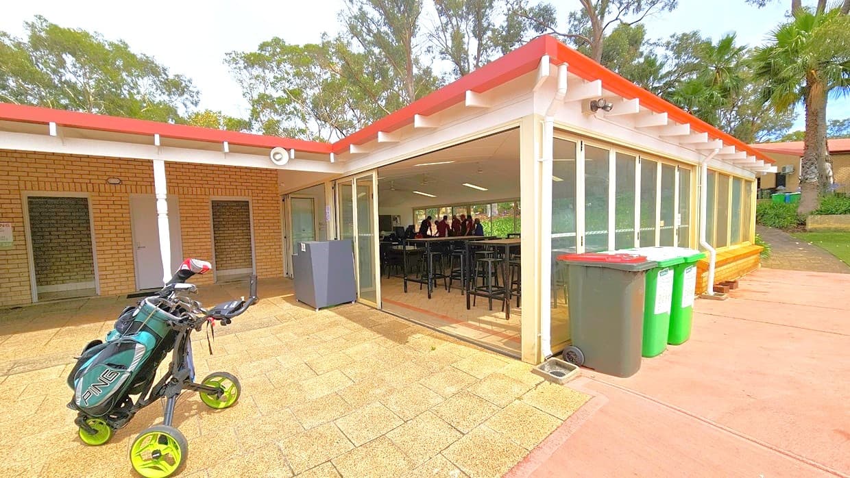 To the left is a brick wall housing the bathrooms. On the right is a room filled with movable chairs and tables. Large rubbish bins are arranged against the glass wall. Behind, there is a cream building and palm trees. A golf trolley is visible in front. The floor has brown tiles, and on the right is a pink concrete floor.