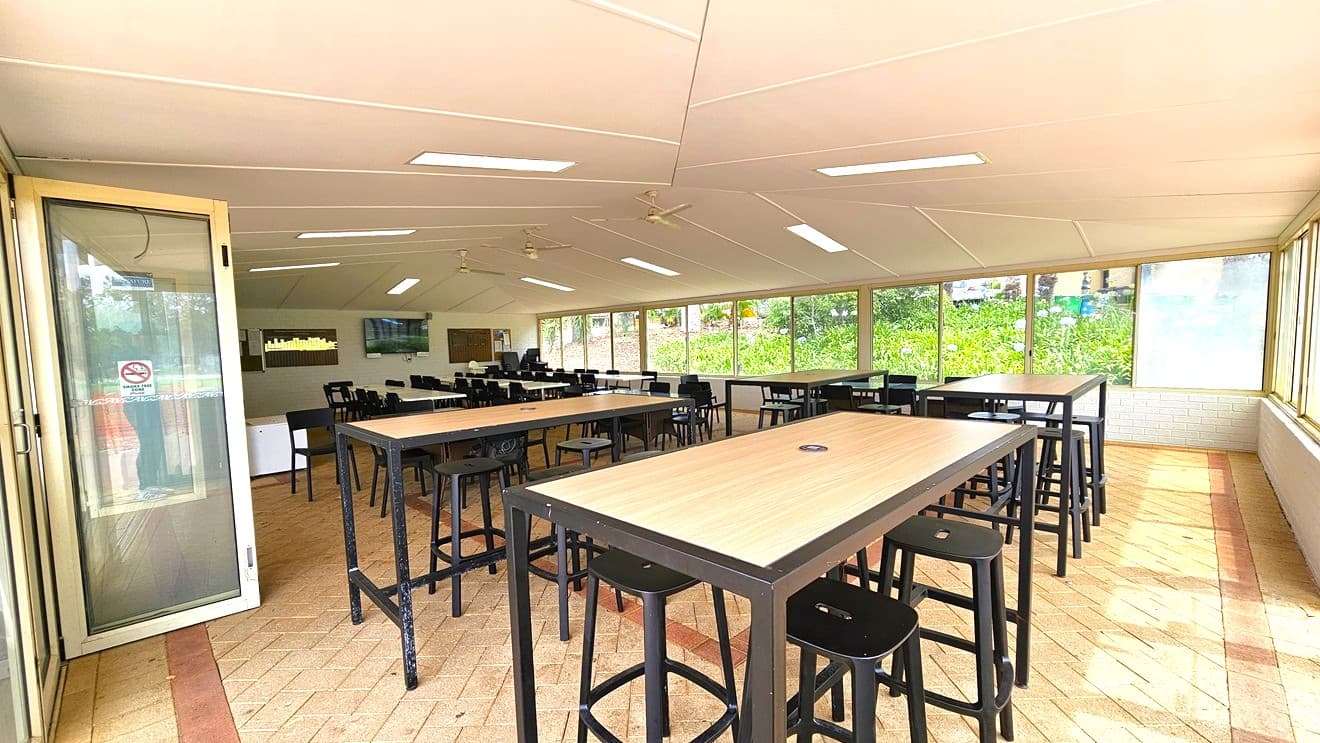 Tables with wooden designs and black stools are arranged. The glass door, bearing a no-smoking sign, opens outward. The left wall features a flatscreen TV. The entire wall on the right is made of glass, allowing a view of the garden outside. The floor has cream tiles with pink accents, and the white ceiling is equipped with fluorescent lights and ceiling fans.