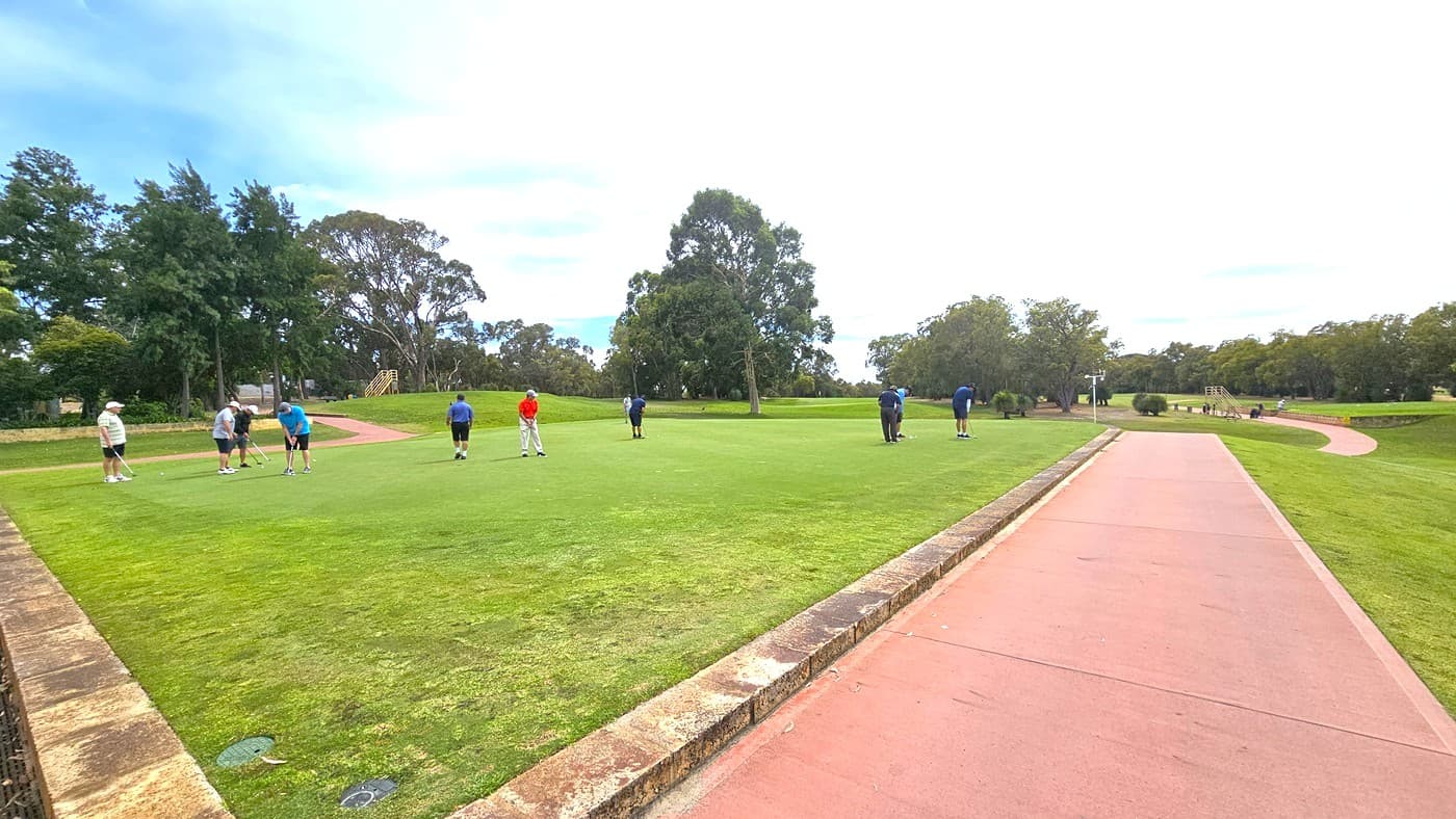 On the right, a pink concrete path leads to the greens. A concrete border separates the path and the putting green, extending across the area. Golf players are currently engaged in a game. The surroundings are characterized by trees and green grass.