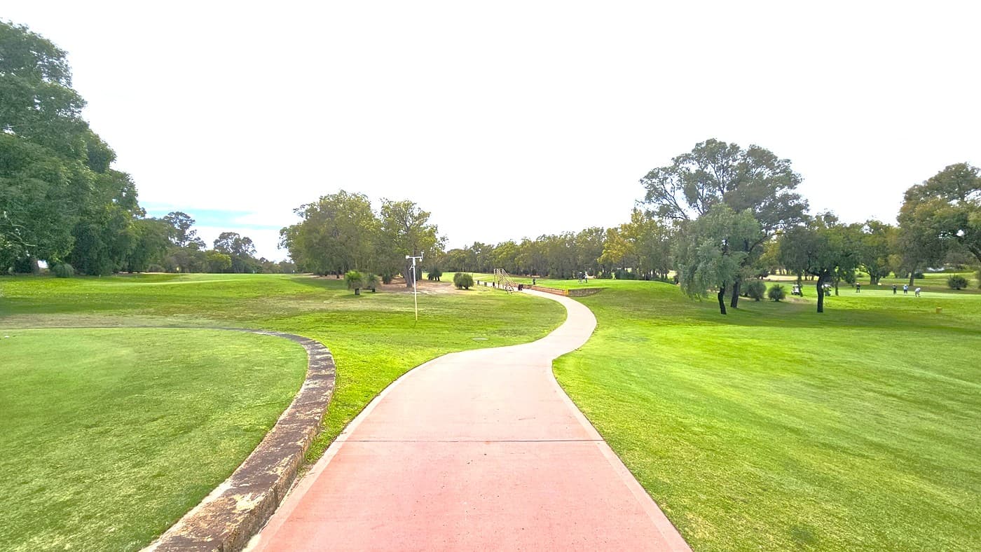 To the right, a pink concrete path descends to the greens, separated by a concrete border that extends across the putting green area. On the right, a sloping hill is covered in green grass. Further ahead, the concrete path continues to the other section of the golf area. The terrain varies in depth and elevation and is surrounded by trees. Golf players are visible.
