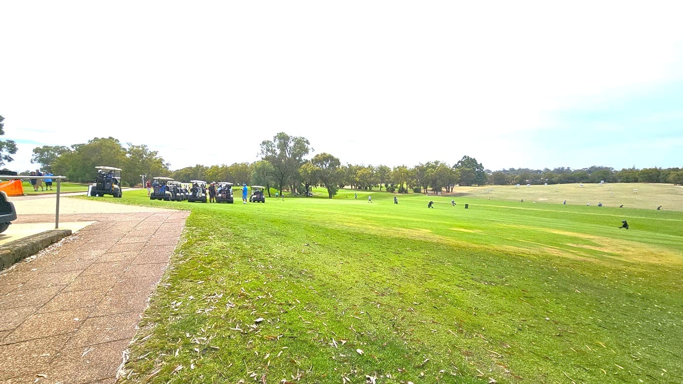 To the left, a concrete path leads to the greens. On the right, a sloping hill is covered with green grass. Golf carts are parked, and golfers are scattered across the green. Further ahead, the concrete path continues to the other section of the golf area, surrounded by trees and hills.