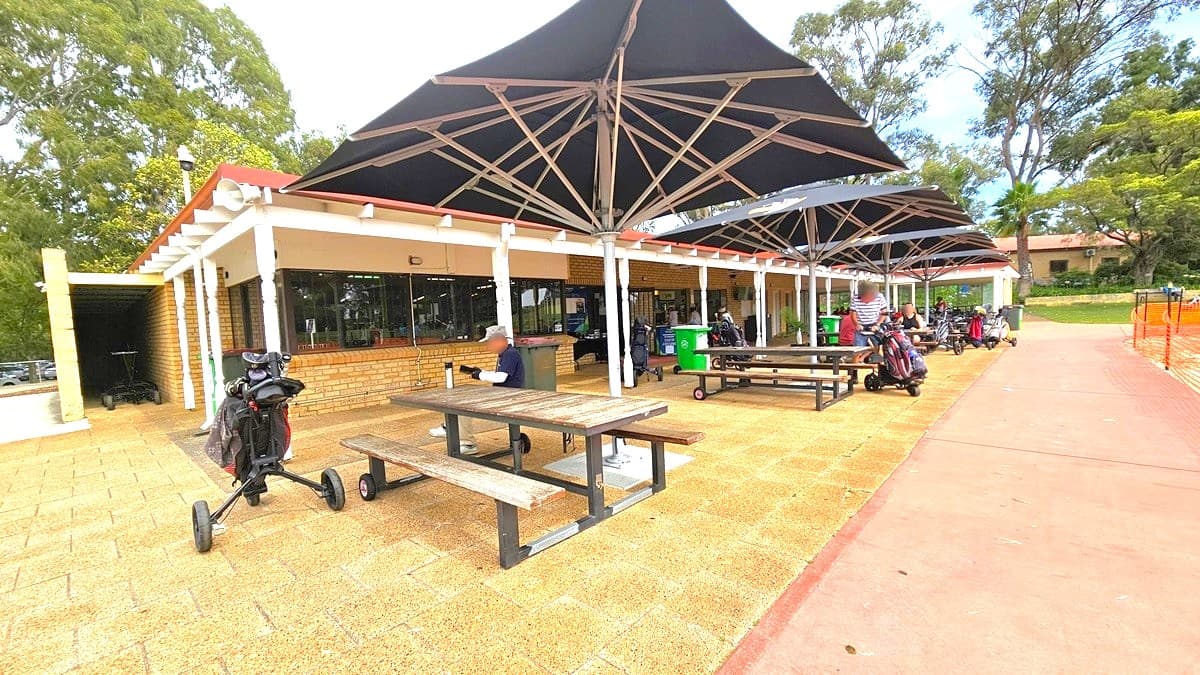 The facility building, constructed with brown bricks and glass. In front, a veranda and a shaded area feature picnic tables and chairs under large black umbrella shades, with people seated underneath. To the left, trolleys are parked. The floor transitions from white concrete to brown tiles and red cement tiles. Tall trees surround the area.