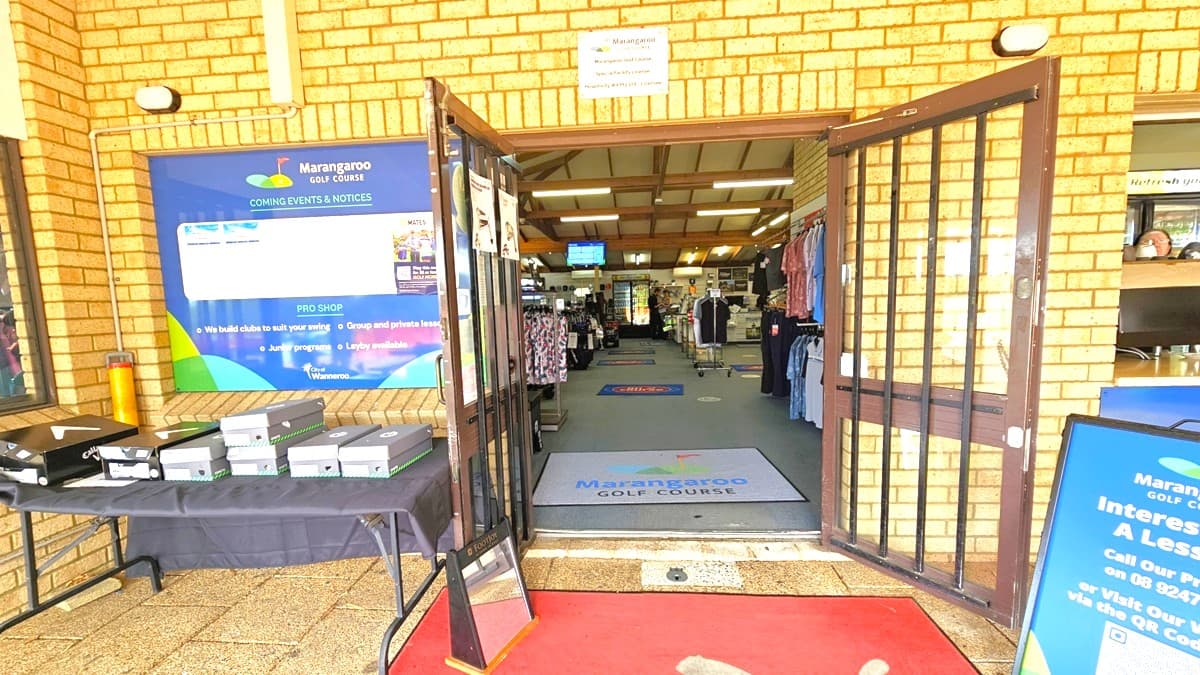 On the left, a table displays golf balls, and to the right stands a free standing banner. The glass door in the middle opens outward. Inside, a large floor mat showcases the facility's name at the doorstep and along the aisle. Retail items are visible, and the building walls feature brown brick, with a large facility banner affixed to the left.