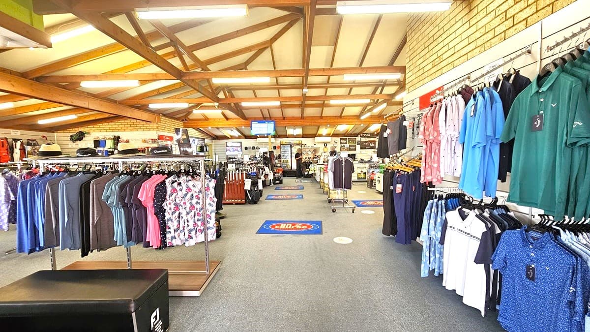 On the right, T-shirts and polo shirts are neatly displayed, while ladies' clothing is on the left. Blue floor mats, showcasing the facility's name, line the floor. Golf bags are prominently featured on the left, with golf clubs displayed near the aisle. Moving forward, the reception counter is visible. The ceiling adopts an industrial aesthetic with exposed timber beams and fluorescent lights. Grey carpets cover the floor, and brown brick walls add to the interior.