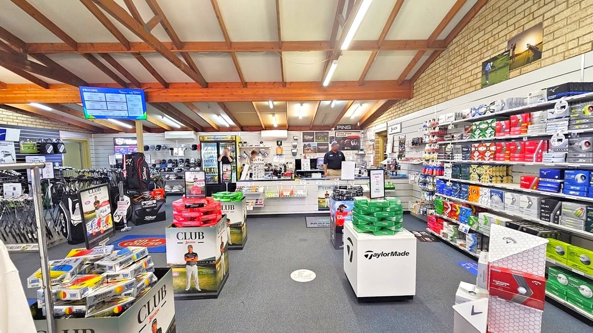 On the right, there is a multi-layered display rack filled with golf balls of various brands, and additional items are displayed in the aisle. On the left, golf bags and Trolley are showcased. A banner displaying pricing hangs from the ceiling. To the right, the wall features golf exhibits. The ceiling adopts an industrial aesthetic with exposed timber beams and fluorescent lights. Grey carpets cover the floor, and brown brick walls add to the interior.