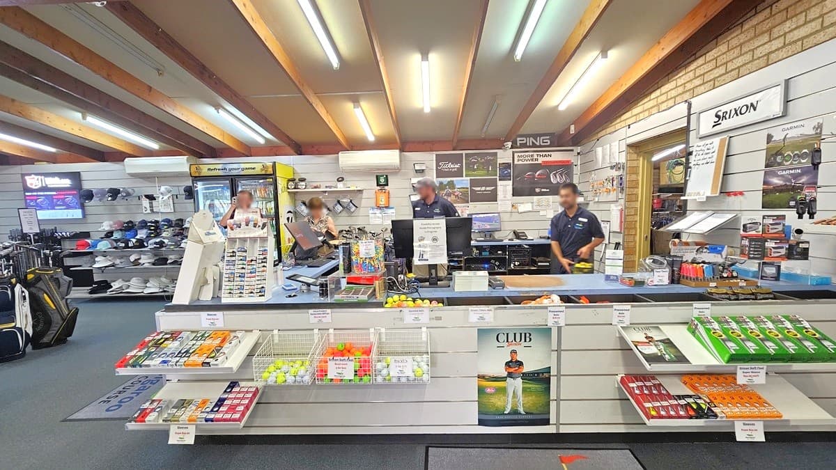 In the middle is the reception counter featuring golf balls on the front. The counter is equipped with computers and POS machines, with staff standing behind the desk. On the left, there is a fridge filled with cold drinks for sale, next to a tiered display rack showcasing caps. Trolley bags are positioned on the left. The ceiling features a wooden plank design with fluorescent lights illuminating the space. The floor has grey carpet, and the wall on the right has brown brick.