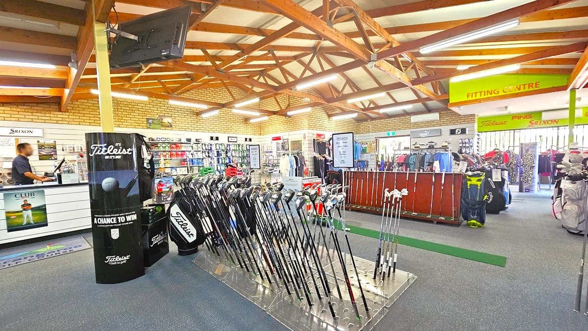 A display rack showcasing golf clubs of various sizes, with a black signage positioned next to it. To the left is the reception counter with staff standing behind, and to the right is another rack displaying clubs, shirts, and bags. Behind is another display rack filled with golf balls. The ceiling features a wooden plank design with fluorescent lights illuminating the space. The floor has grey carpet, and the wall on the right has brown brick.