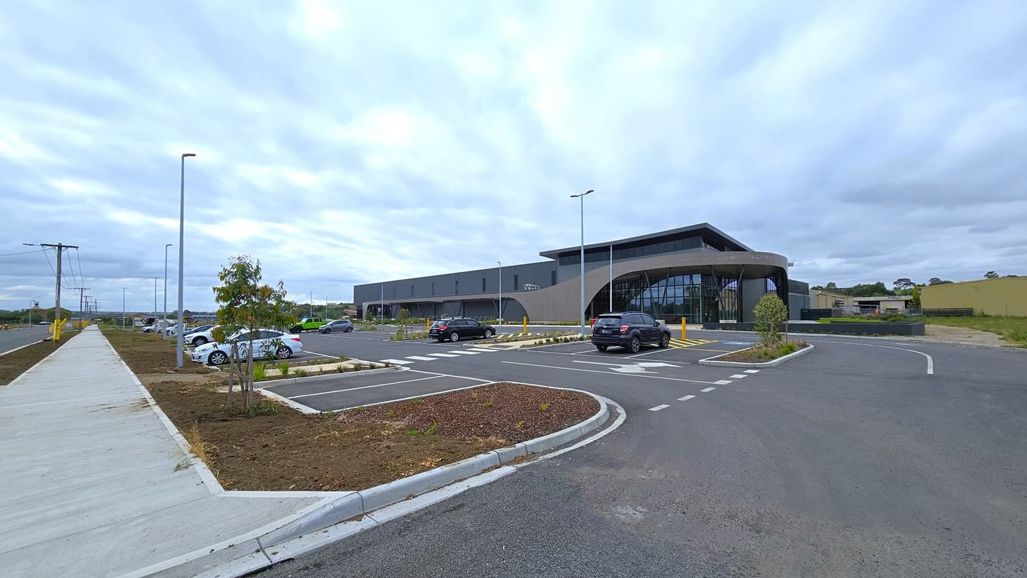 A car park with parked vehicles. Concrete pavement separates it from the road. Accessible parking is marked with signs and yellow bollards. A concrete plant box has plants and small trees. Next to it is a modern facility building, with a grass area to the right.