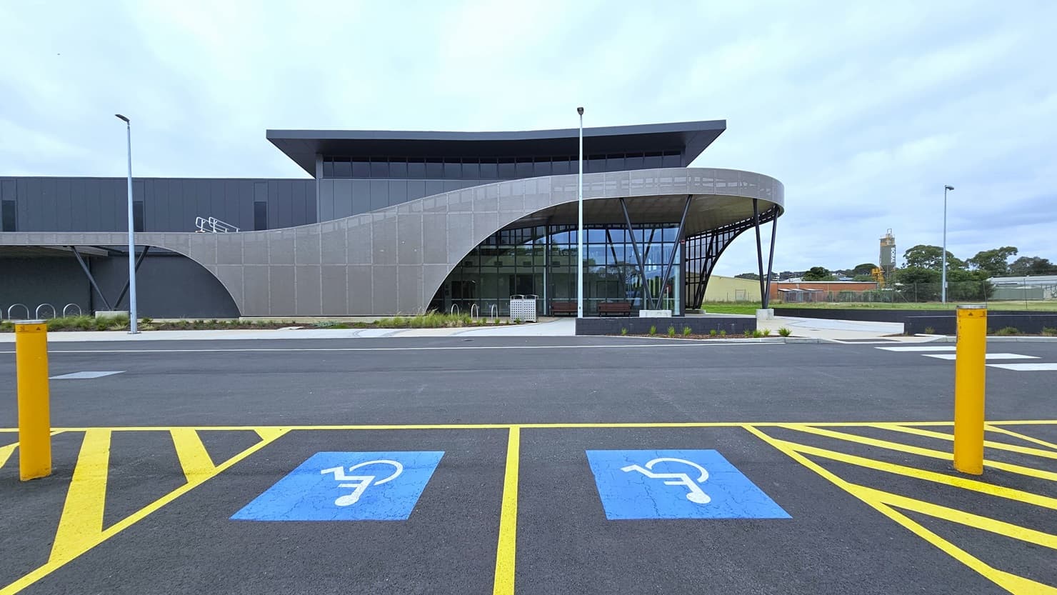 Accessible car parking spaces are marked with signs and yellow bollards. Adjacent is a modern facility building made of concrete, glass, and beams. A grass area lies to the left, bordered by tall lamp posts along the road. Bike racks are positioned at the sides of the building. A concrete plant box hosts plants and small trees.