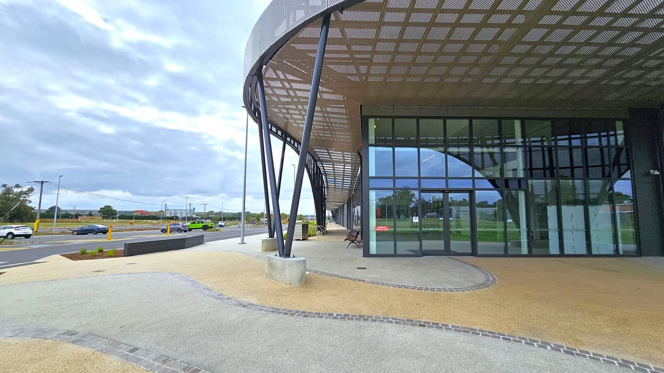 A concrete pavement adorns the concourse, with vehicle parking on the far left. In the middle stands a modern facility building of concrete and glass, featuring a large circular shade with steel mesh. The entrance boasts a floor-to-ceiling glass wall with an automatic door. Chairs and bike racks are visible on the left side.