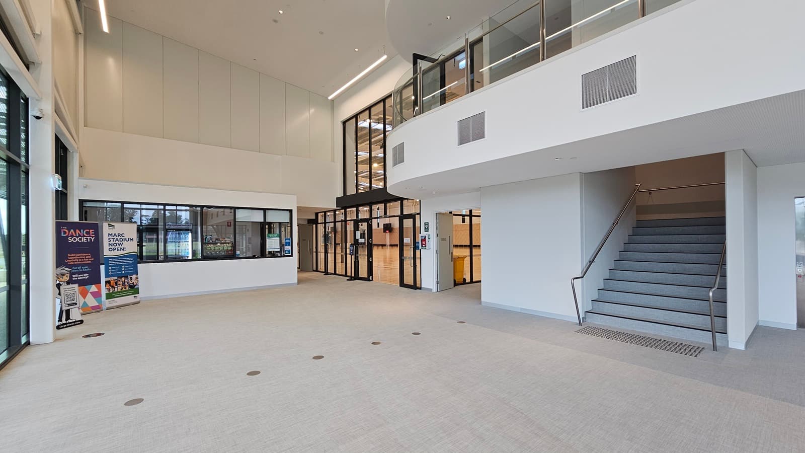 To the right is a staircase with handrails and truncated domes. Inside, a high ceiling accommodates a mezzanine. Straight ahead is the reception desk. Retractable banners are positioned on the left, near the glass window. To the right of the reception is the stadium, secured with glass walls. The floor boasts a firm vinyl finish.