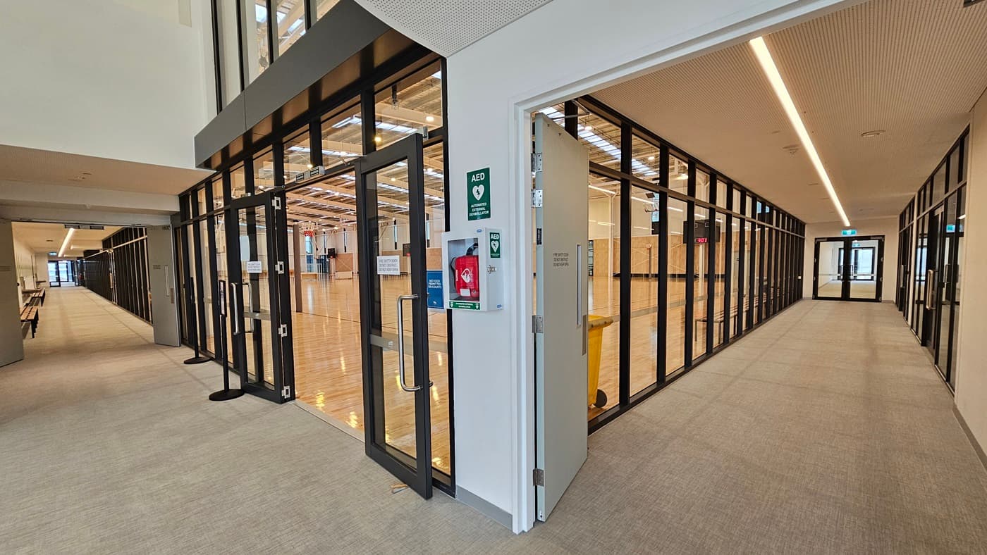 The stadium entrance features a manual outward-opening glass door with wall signage, including a defibrillator. To the right, a corridor leads to toilets, lockers, and change rooms. On the left, another corridor leads to bathrooms, Change Places, and the Function Room. Bench chairs and drinking fountains are mounted on the wall. The wall is adorned with foliage decoration, while the ceiling is white. The floor is firm vinyl.