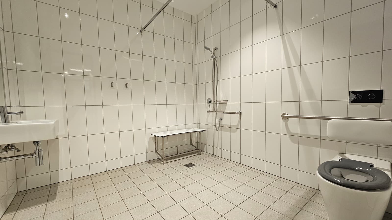 Inside the accessible toilet, a wash basin is positioned to the left. Next to it is a shower with hand grabs and a foldable bench chair. To the right is a toilet bowl with hand grabs. White tiles cover the entire floor and wall. Above, a placeholder for a curtain hangs in place.