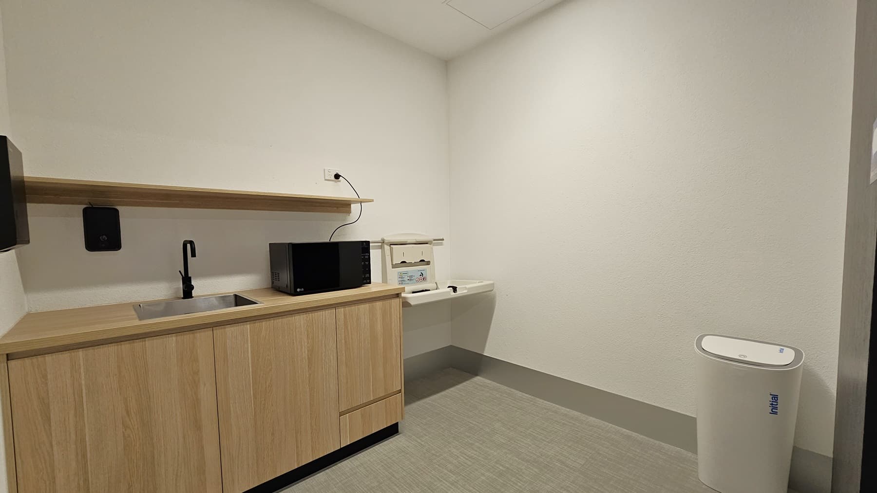 On the left, there's a wash basin with a wooden cabinet and ledge. A microwave oven sits on top. Soap and tissue dispensers are fixed to the wall. A white rubbish bin stands on the right. The ceiling is white, and the floor is firm vinyl.