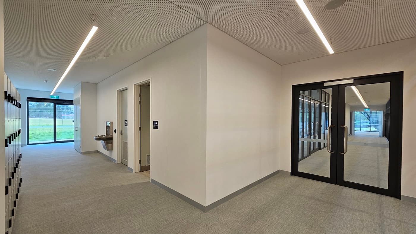 On the left, there's a 4-tier locker area. In the middle, an accessible toilet is marked with a gender-access sign. A drinking faucet is fixed to the wall. At the far end, a glass wall offers an outside view. To the right, a manual glass door. The wall and ceiling are white with LED lights. The floor has firm vinyl.