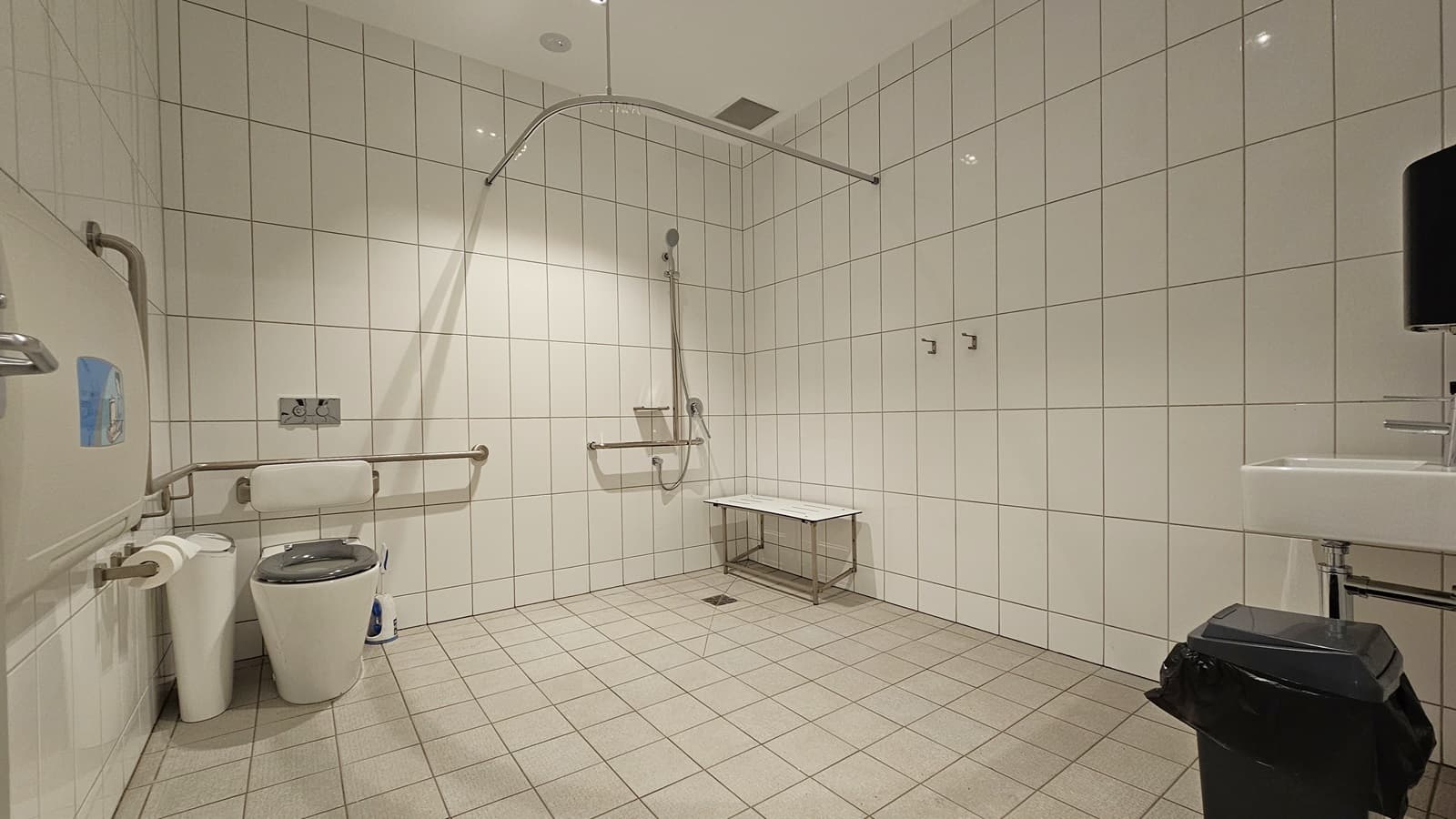 Inside the accessible toilet, a wash basin is on the right with a black rubbish bin nearby. In the middle, a toilet bowl with hand grabs and a shower with a foldable bench chair underneath. On the left, a foldable change table is mounted to the wall next to a rubbish bin and tissue dispenser. White tiles cover the floor and wall, with a curtain holder above.