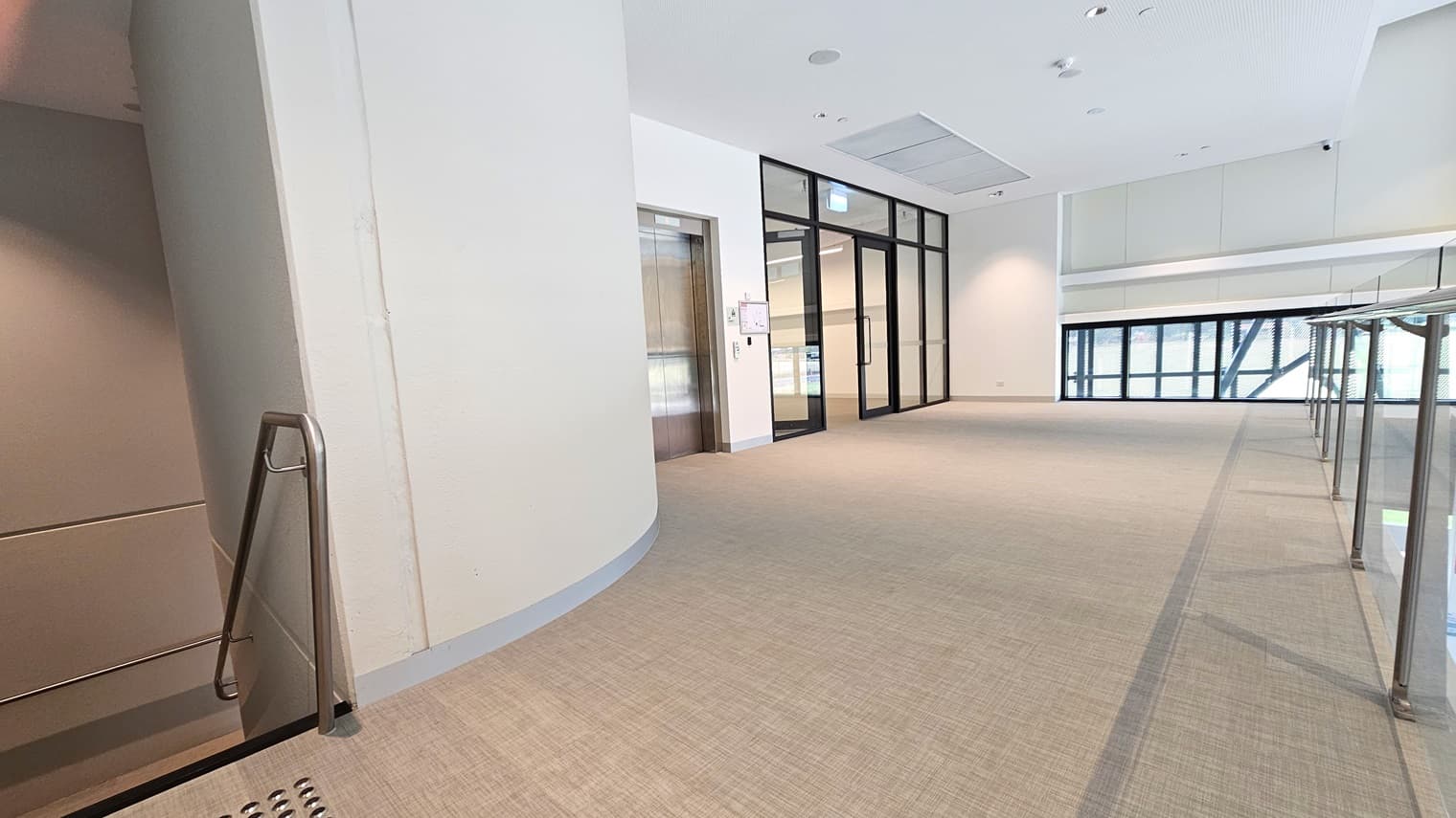 The mezzanine floor includes a lift on the right and stairs with handrails and truncated domes. A view of the lobby below is visible, along with retractable banners in front of the reception. Far ahead is a glass door that opens outward. The floor has vinyl tiles, and the wall and ceiling are all white with a porous design and LED lights.