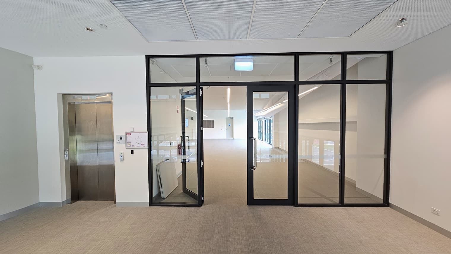To the left is a lift. Next to it is a manual inward-opening glass door leading to the Function Room, surrounded by glass. On the right is another lift and a manual glass door. A site map is affixed to the wall. A foldable table rests against the left wall. The floor has firm vinyl, and the walls and ceiling are all white.