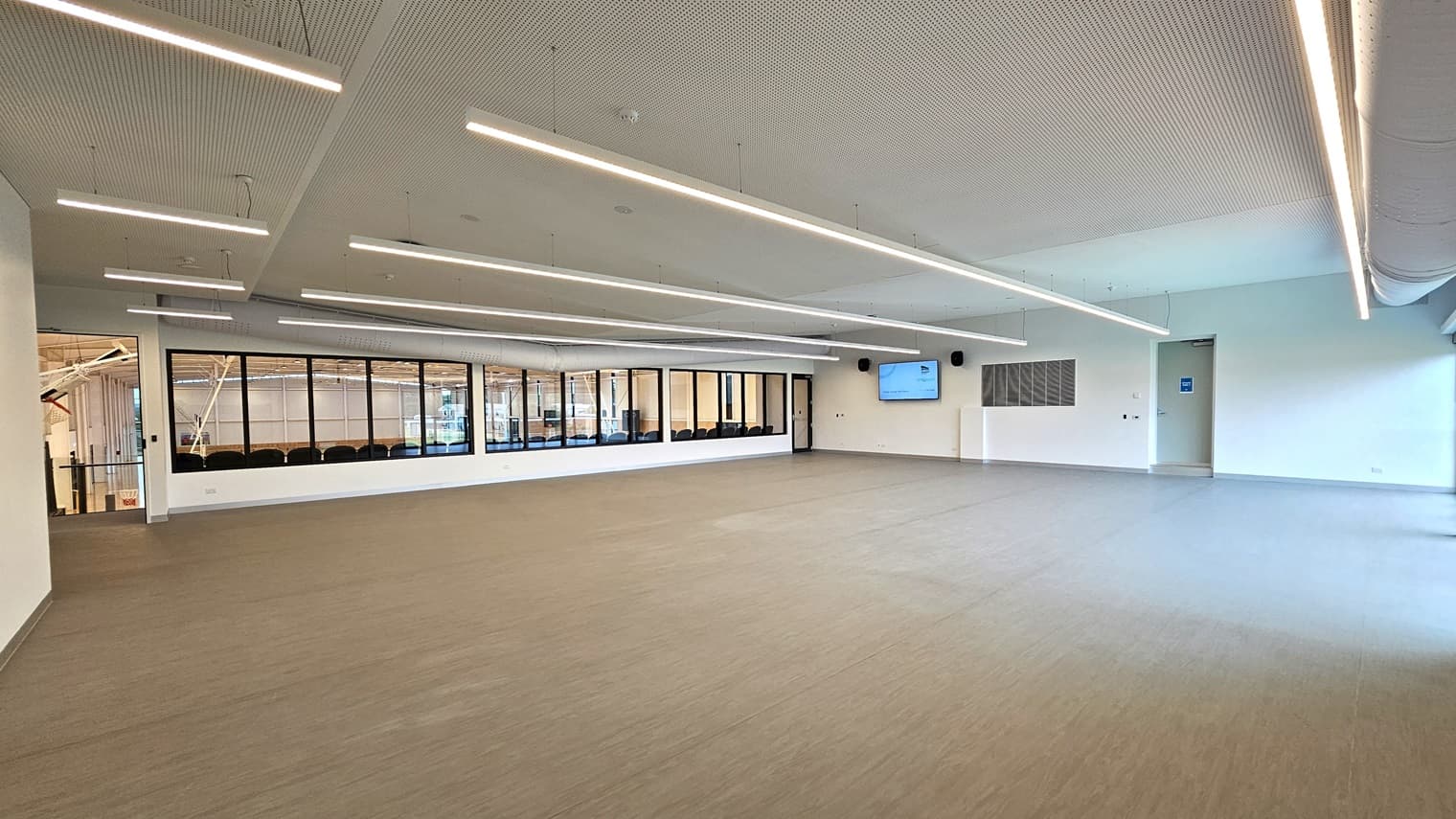 The Function Room is a spacious area with vinyl flooring. A large flatscreen TV is mounted on one wall. Three doors provide access, including one leading to the stadium's spectator area. The walls are white, and the ceiling features a white porous design with fluorescent lighting.