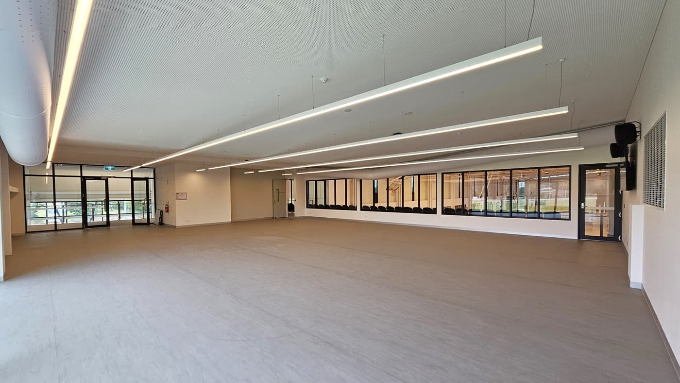 The Function Room is a spacious area with vinyl flooring. A large flat-screen TV and audio speakers are mounted on the right wall. Three doors provide access, two leading to the stadium's spectator area and another on the left leading to the lift. The walls are white, and the ceiling features a white porous design with fluorescent lighting and exposed large air conditioning vents.