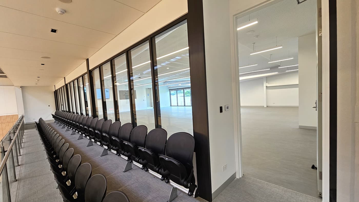 To the right is the Function Room, a spacious area with vinyl flooring. A large flat-screen TV and audio speakers are mounted on the right wall. Adjoining is the stadium's spectator area with chairs arranged for seating. A glass railing secures the area. The walls are white, and the ceiling features a white porous design with fluorescent lighting.