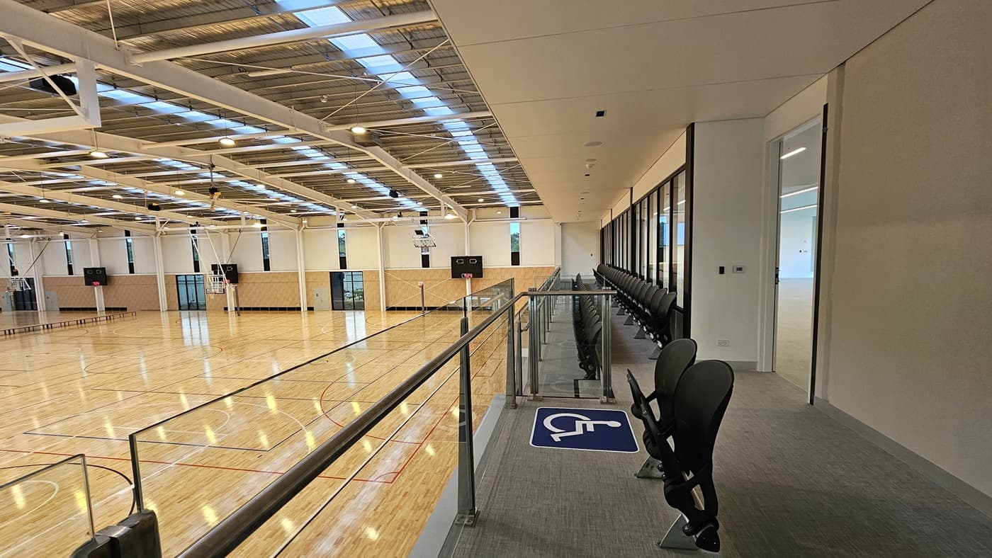 To the right is the Function Room, a spacious area with vinyl flooring. Adjoining is the stadium's spectator area with two rows of folded chairs for seating, including accessible seating. A glass railing secures the area. To the left is the stadium, featuring timber floors and an industrial ceiling with exposed beams, skylights, and aluminum insulation.
