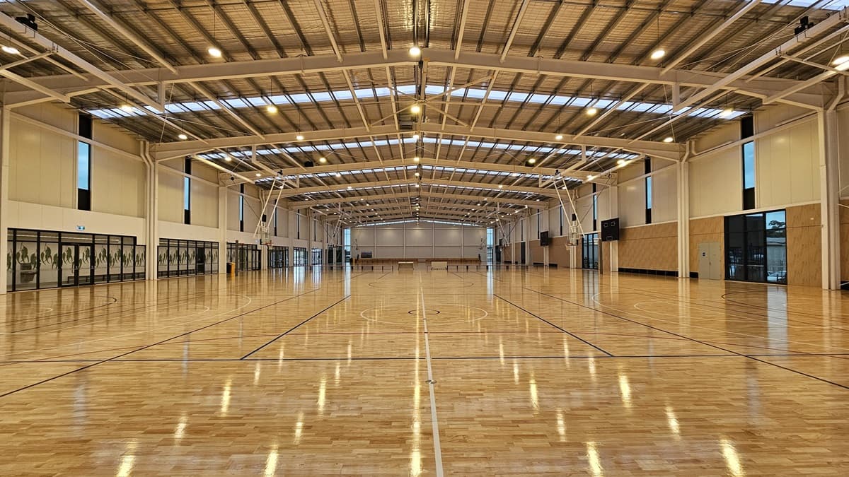 The stadium is a spacious area with polished timber flooring marked for various ball games. The ceiling has an industrial aesthetic with exposed beams, aluminum insulation, and ample skylights. On the left, a mezzanine offers seating for spectators, surrounded by glass.