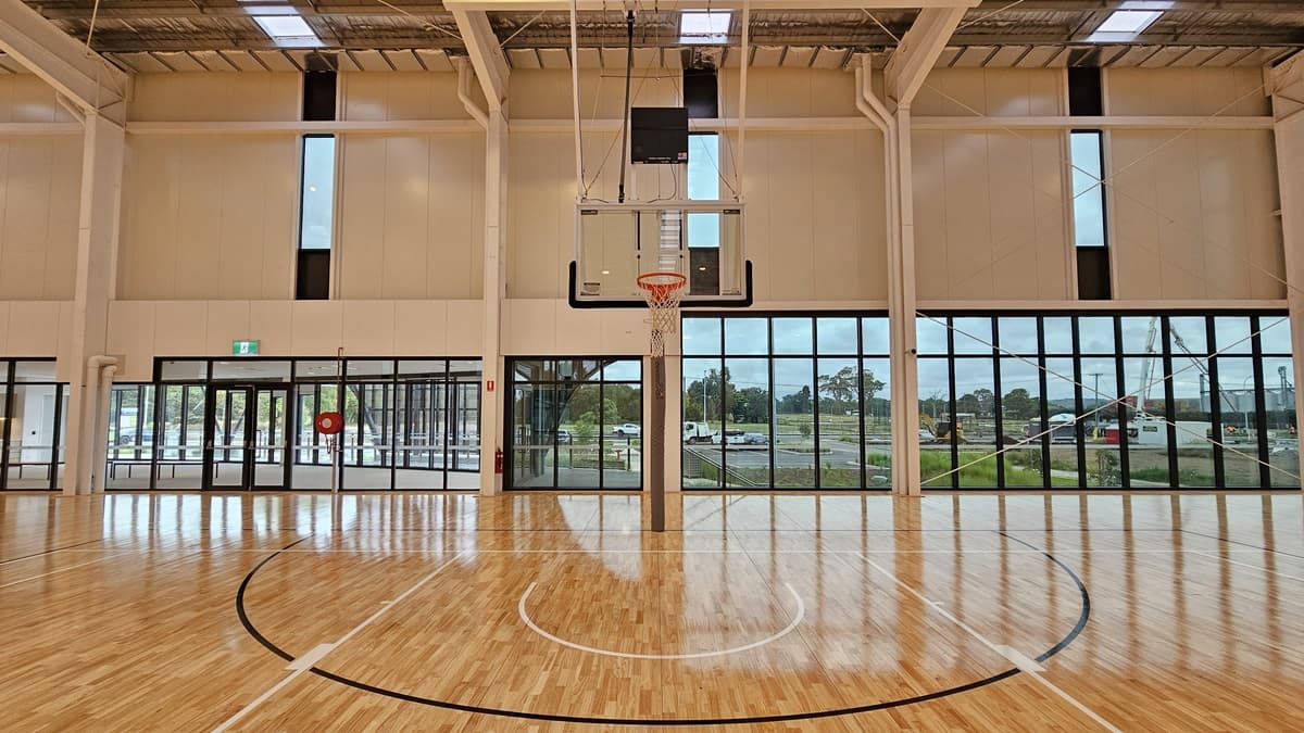 A spacious seating area overlooks the stadium with bench chairs positioned in the middle. The stadium, surrounded by glass, features timber flooring. At the far end is a glass door marked with an "Exit" sign. The floor is firm vinyl, and the ceiling has a white porous design with fluorescent lighting.