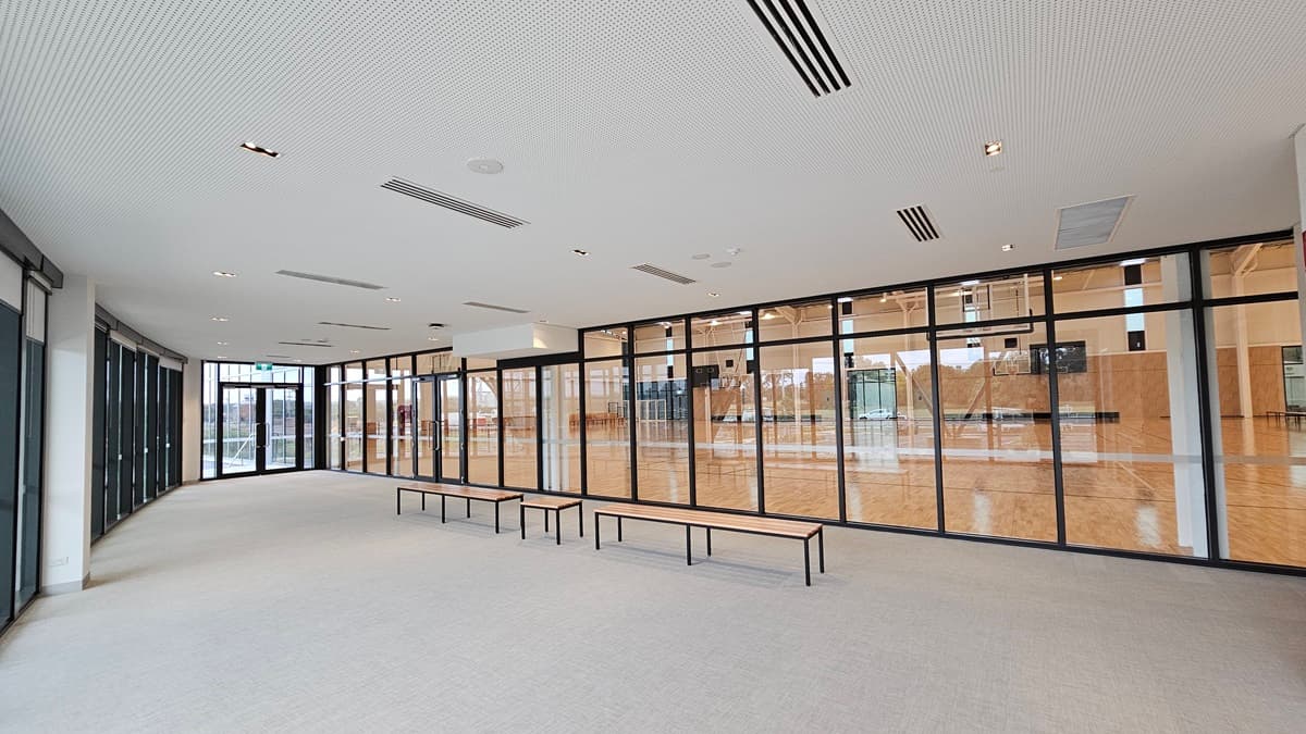 A spacious seating area overlooks the stadium with bench chairs positioned in the middle. To the left, there is a wash area with a small fridge. The wall is made of glass, providing a view of the scenery outside. At the far right is a glass door marked with an "Exit" sign. The floor is firm vinyl, and the ceiling has a white porous design with fluorescent lighting.