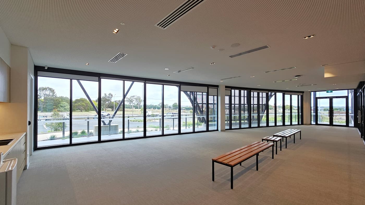 The mezzanine area features seats for spectators, including accessible seating, each marked. A stainless steel glass railing secures the area. From this view, the court below is visible, featuring polished timber floors and an industrial-look ceiling with exposed beams, aluminum insulation, and skylights.