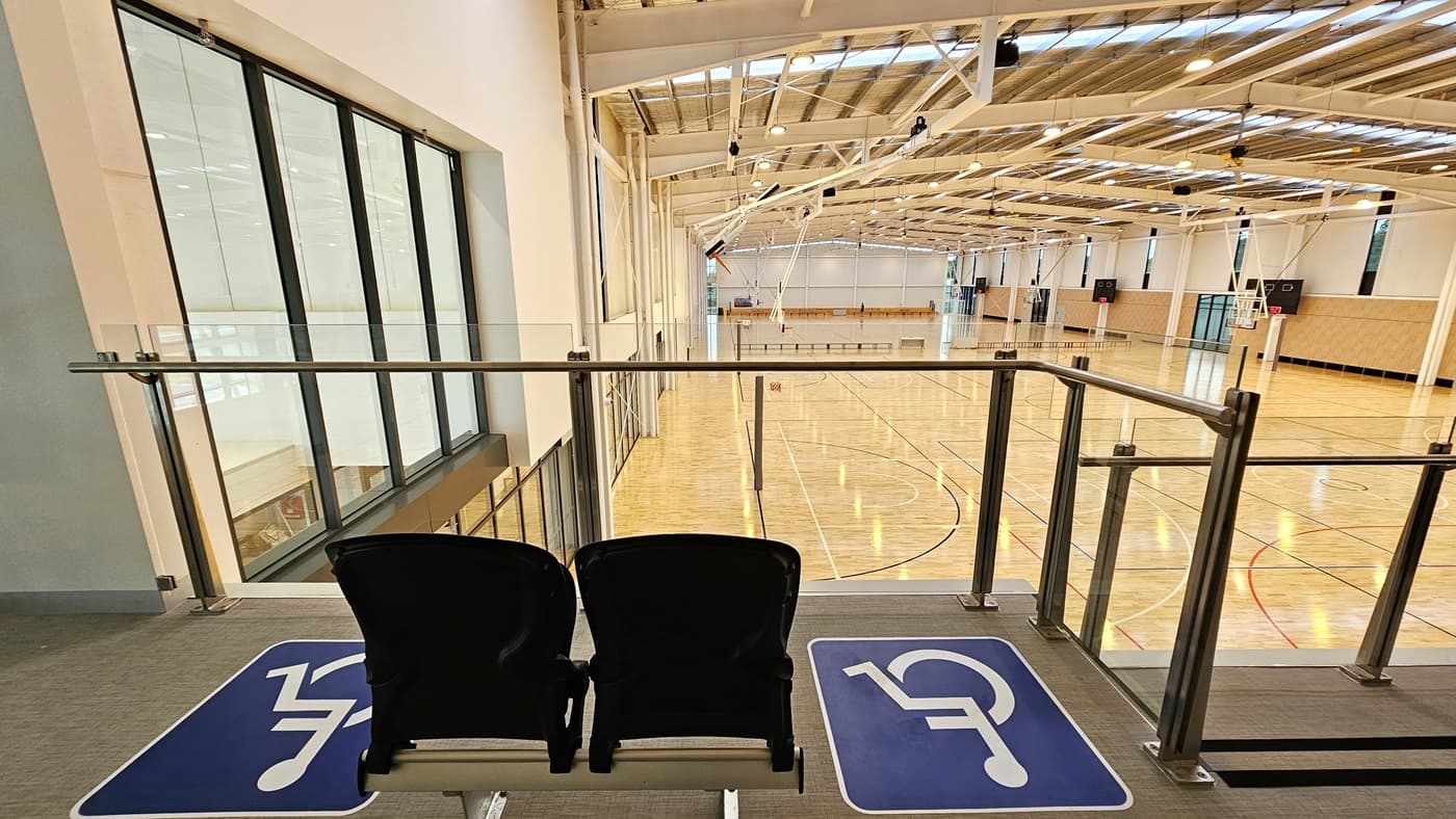 The mezzanine area has two rows of seats for spectators, including accessible seating, each marked. A stainless steel glass railing secures the area. From here, the court below is visible, with polished timber floors and an industrial-style ceiling featuring exposed beams, aluminum insulation, and skylights.