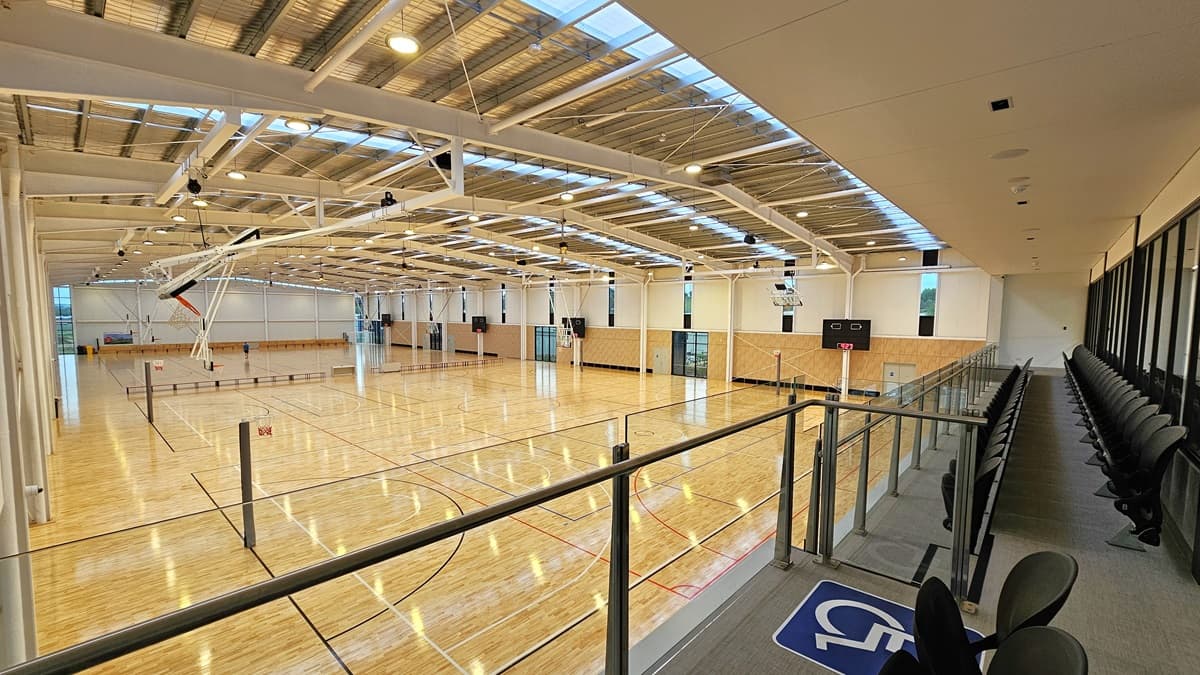 To the right is the stadium's spectator area with two rows of folded chairs for seating. A glass railing secures the area. To the left is the stadium, featuring timber floors marked for various ball games and an industrial ceiling with exposed beams, skylights, and aluminum insulation.
