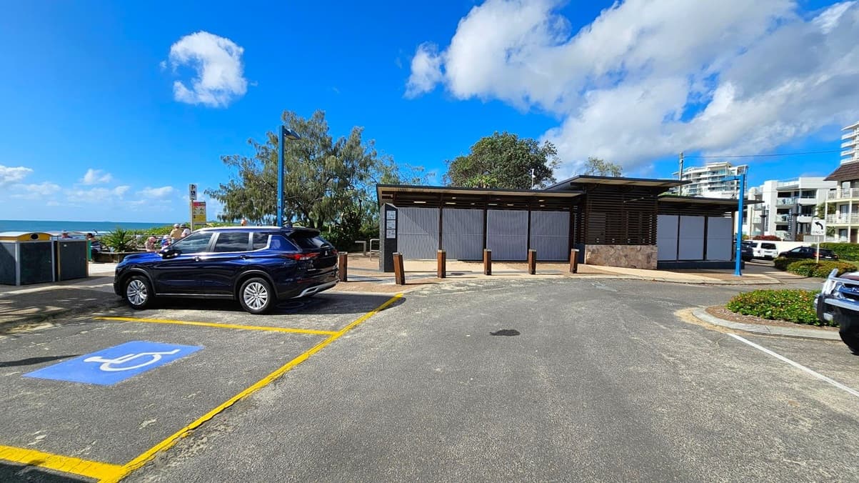 A car is parked in a designated disabled parking space on the left, marked with a blue and white symbol. In the centre, there is a small building with a corrugated metal facade and a flat roof. Trees and shrubs are visible behind the building. On the right, there are more parked cars and a few multi-storey buildings in the background. The ground is paved with asphalt, and there are some wooden bollards near the building.