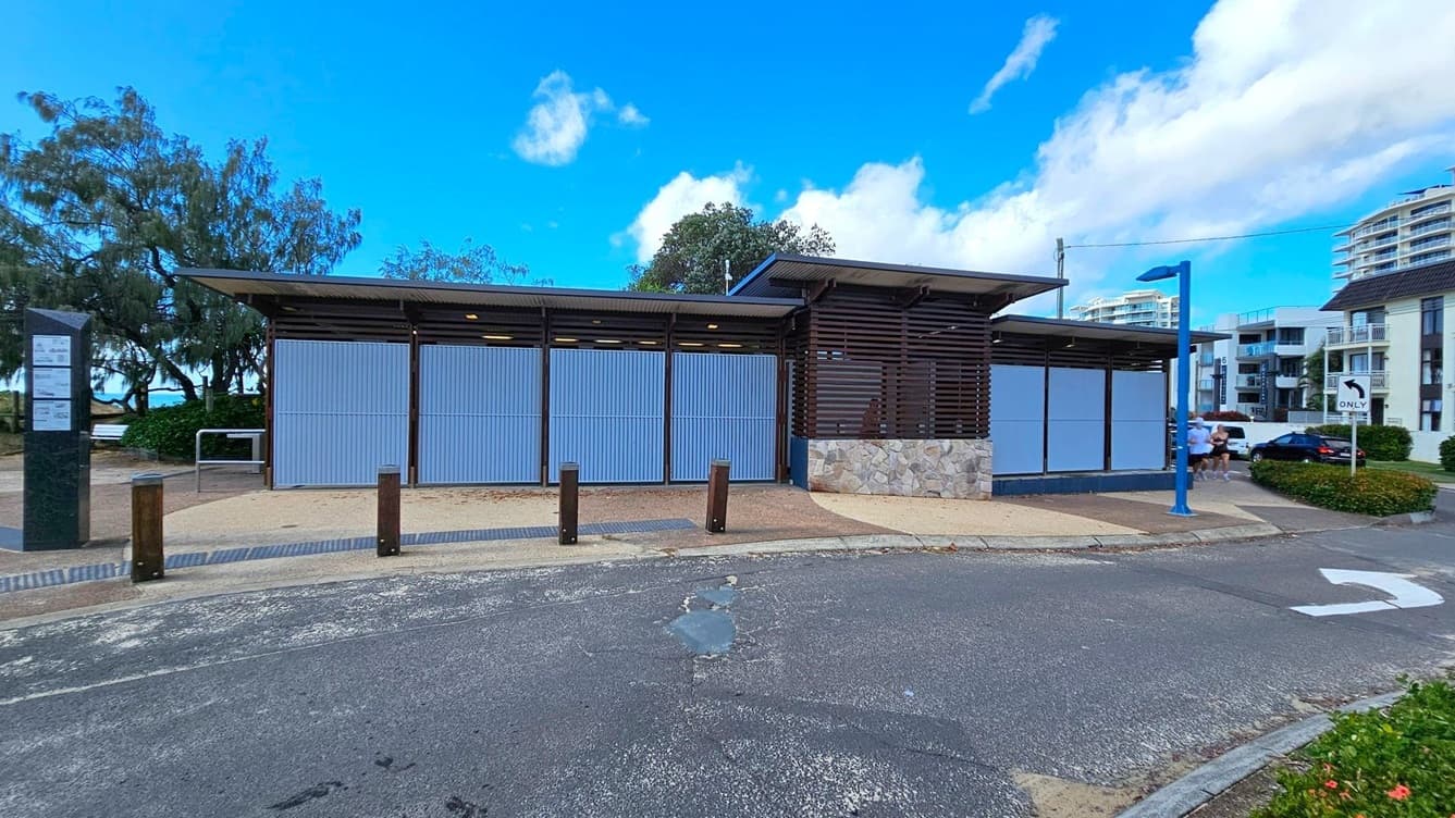 A modern public restroom facility with a flat roof and light blue corrugated panels. The structure has a stone base and dark horizontal slats on the right. In the foreground, there is a paved area with bollards and a road with a painted arrow. On the left, trees and a glimpse of the sea are visible. On the right, a blue street lamp and several multi-storey buildings are in the background. A few people are walking near the building.