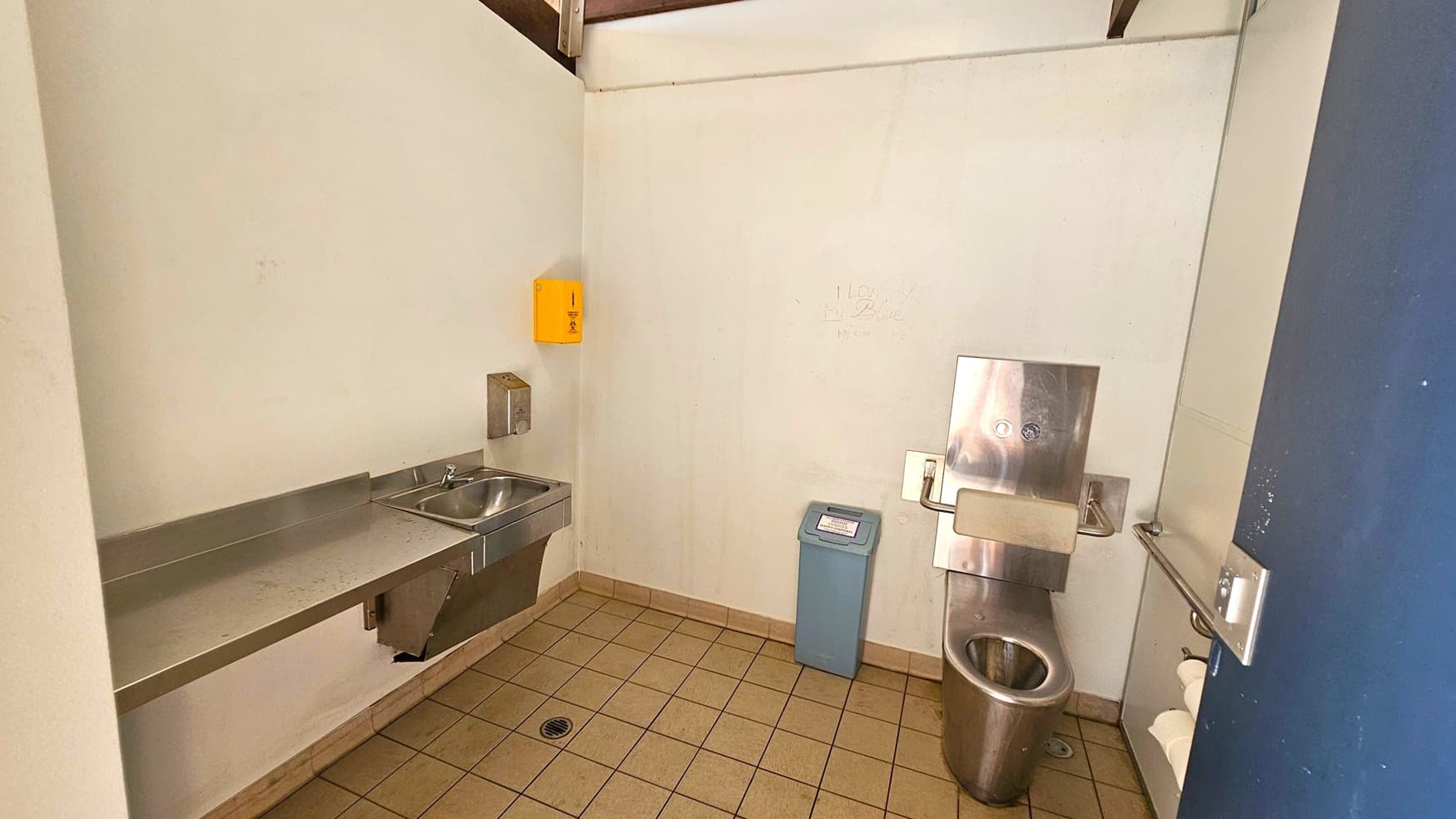 A public restroom with a stainless steel toilet on the right, featuring a mounted flush panel. Next to it is a blue waste bin. On the left, a stainless steel sink with a soap dispenser above it. A yellow box is mounted on the wall above the sink. The floor is tiled with a drain in the centre, and the walls are plain white. A metal bar is attached to the wall near the toilet.