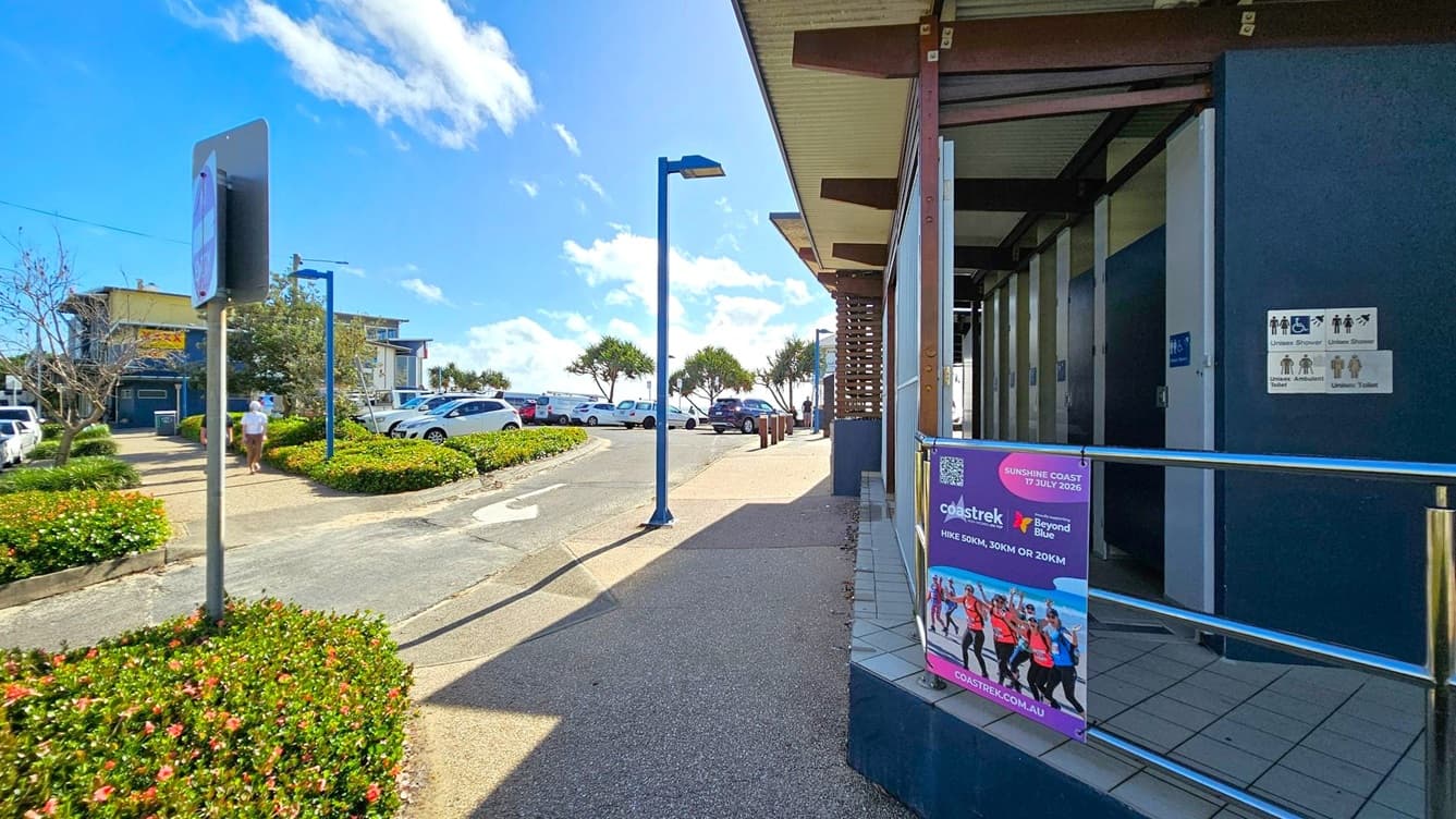 On the right, there is a public restroom facility with signs indicating accessible toilets and showers. A poster promoting a hiking event is attached to the railing. The left side features a pathway lined with flowering bushes and a pedestrian walking. In the background, there are parked cars and trees near the shoreline. The pavement is a mix of concrete and tiles, and street lamps are visible along the path.