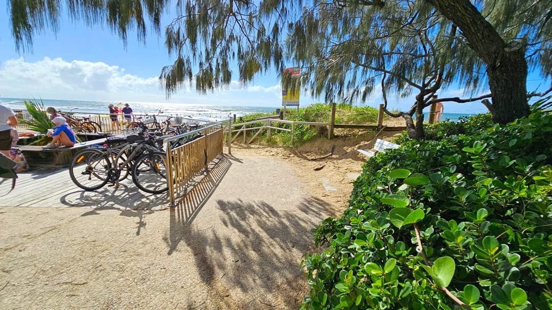 A sandy path leads to a beach area with a wooden fence on the right and lush green bushes. On the left, several bicycles are parked near a wooden deck where people are sitting and enjoying the view. In the background, the ocean is visible with waves and a few people near the water. Overhead, tree branches with long leaves provide some shade.