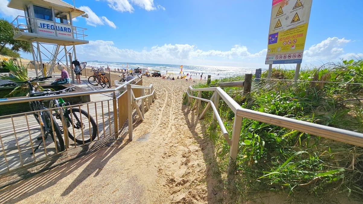 A sandy path leads to a beach with people and flags in the distance. On the left, a lifeguard tower stands on a wooden deck with bicycles parked nearby. A person sits on the deck, and another stands with a surfboard. On the right, a large sign with warnings and information is visible, surrounded by green vegetation. The ocean is in the background, with waves and a bright horizon.