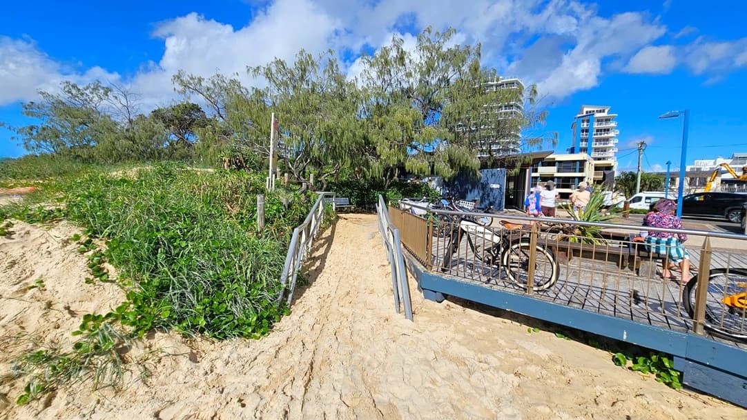 A sandy path leads from the beach to a paved area. On the left, a person is sunbathing on the sand surrounded by greenery. In the centre, trees line the path, and a railing runs along the right side. In the background, several people are walking near parked bicycles and a bench. Tall buildings and a few parked cars are visible further back. The sky is blue with some clouds.
