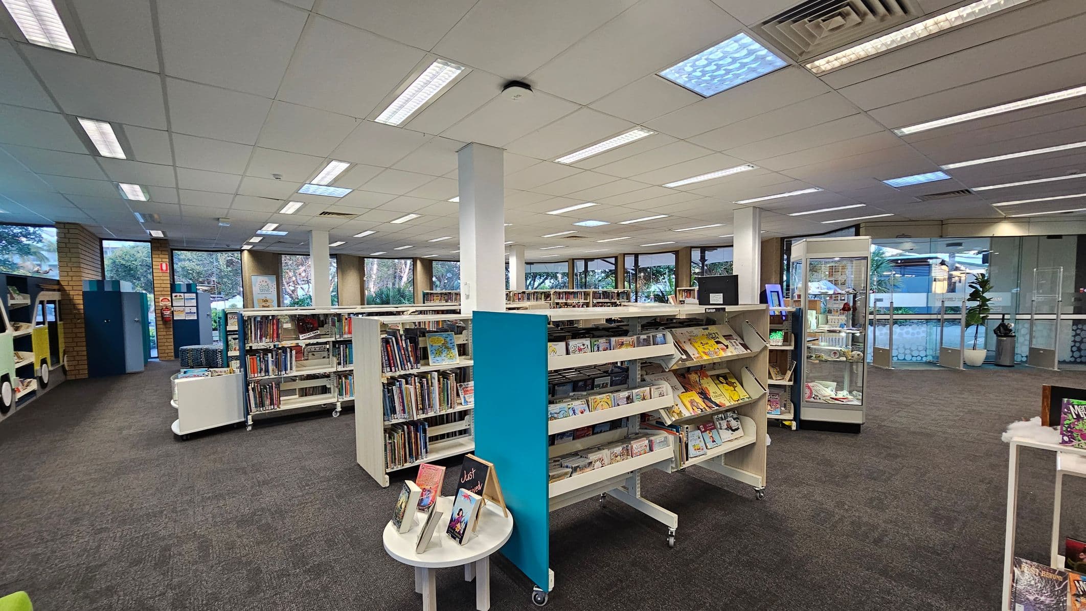 A library interior with grey carpeted flooring and a white panelled ceiling with fluorescent lights. In the centre, there are several white bookshelves filled with books and magazines. On the left, a colourful book display shaped like a vehicle. On the right, a glass display case and a potted plant near the entrance. Large windows in the background provide natural light and a view of greenery outside.