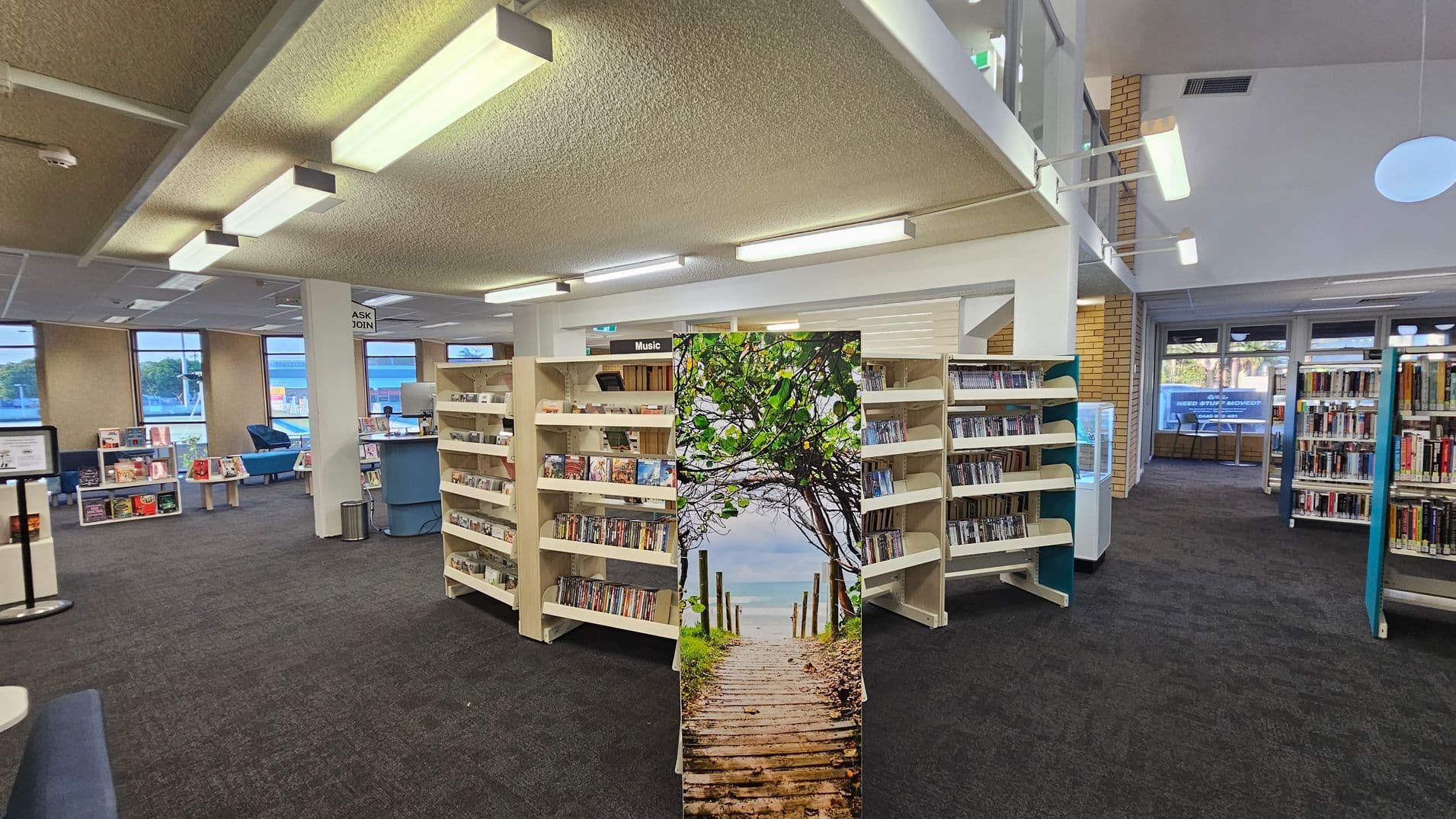 A library interior with a dark carpeted floor and a textured ceiling with fluorescent lights. In the centre, there are bookshelves filled with DVDs and CDs, labelled "Music". A decorative panel with an image of a wooden path leading to the sea is in front of the shelves. To the left, there are windows, a seating area with blue chairs, and a small desk. On the right, more bookshelves are visible, and large windows in the background let in natural light.
