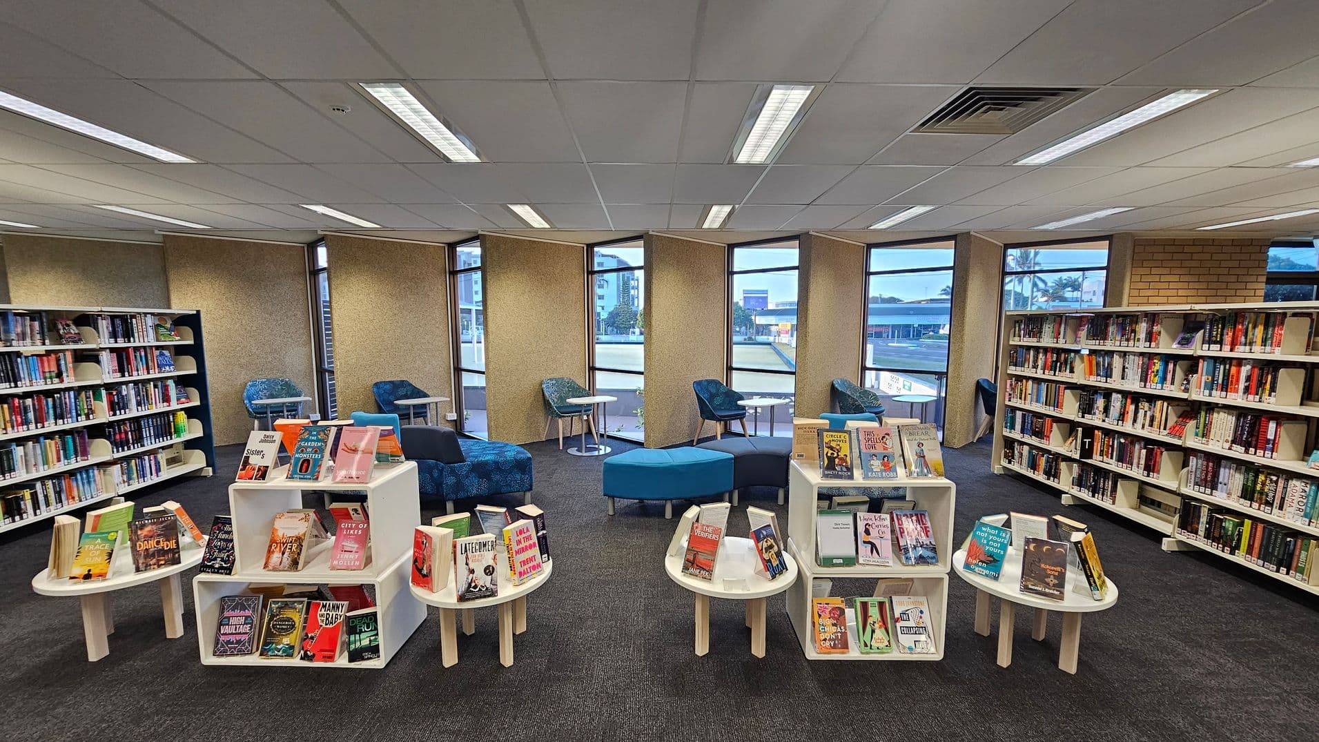A library interior with bookshelves lining the left and right walls, filled with books. In the centre, there are white display tables showcasing various books. The background features large windows with views of buildings outside. Blue upholstered chairs and round tables are arranged near the windows. The floor is carpeted in dark grey, and the ceiling has white panels with fluorescent lighting.
