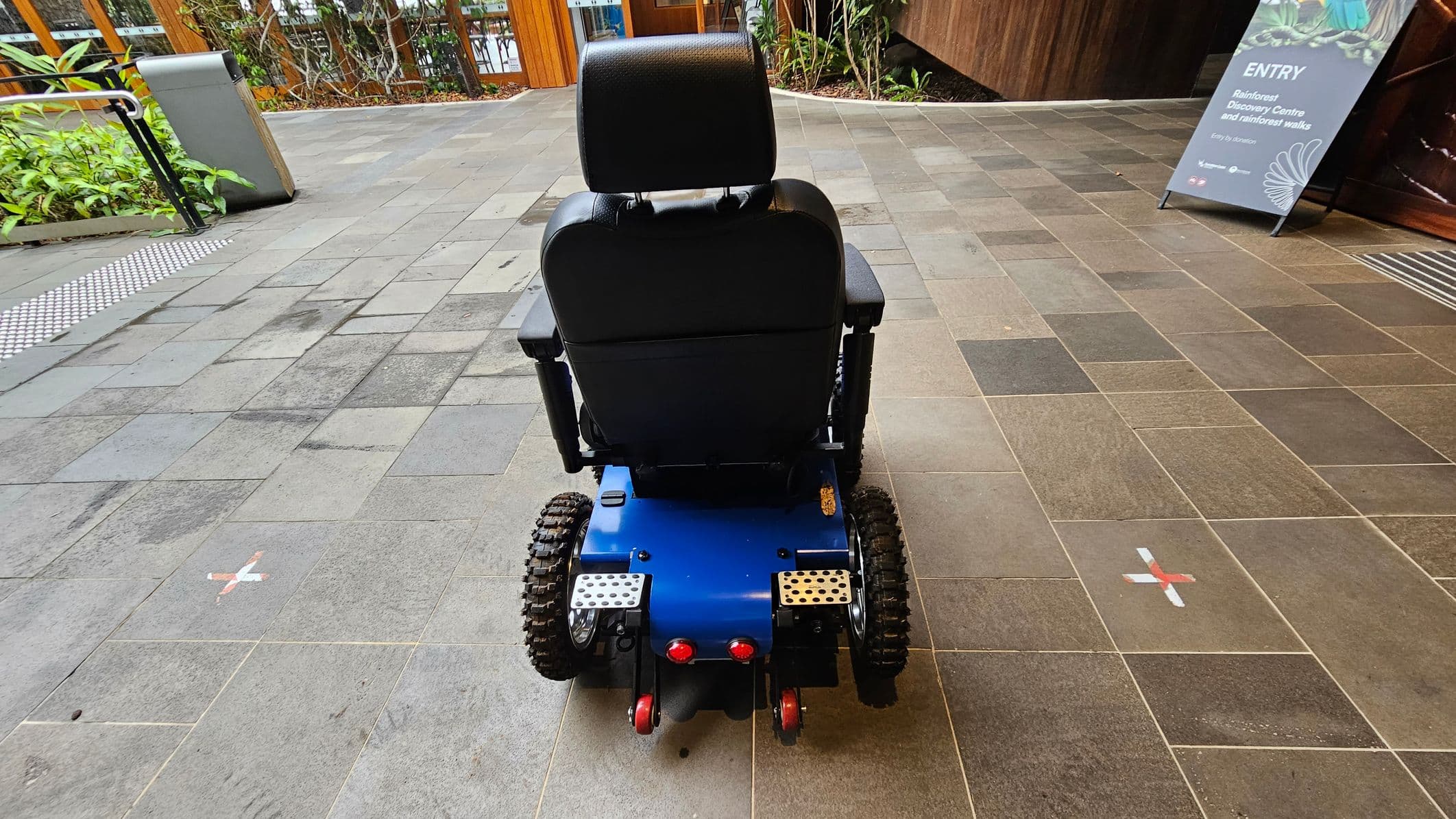 A motorised wheelchair with large, rugged tyres and a blue base is positioned on a tiled floor. To the left, there's a café with glass windows and a wooden sign. In the background, a wooden staircase with plants at its base leads upwards. On the right, a large wooden door with intricate carvings stands open, leading into a building with stone and wood walls.