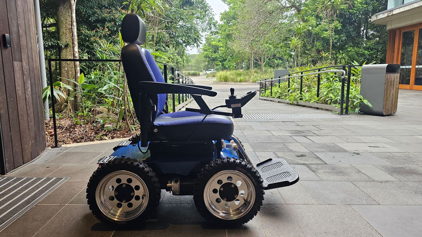 A blue electric wheelchair with large, rugged tyres is positioned on a tiled floor. It has a joystick control on the right armrest. To the left, there's a wooden wall and some foliage. On the right, a ramp with a metal railing leads away, bordered by green plants. A grey bin is near the ramp. In the background, there are trees and a path.