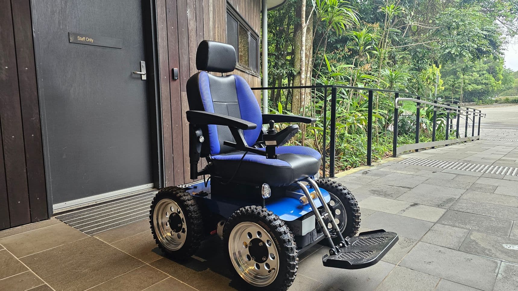 A blue and black electric wheelchair with large, rugged tyres is positioned on a tiled floor. To the left, there is a dark wooden door with a "Staff Only" sign. In the background, lush green foliage lines a pathway with a metal railing. The area is outdoors, with a mix of grey and green tiles on the ground.