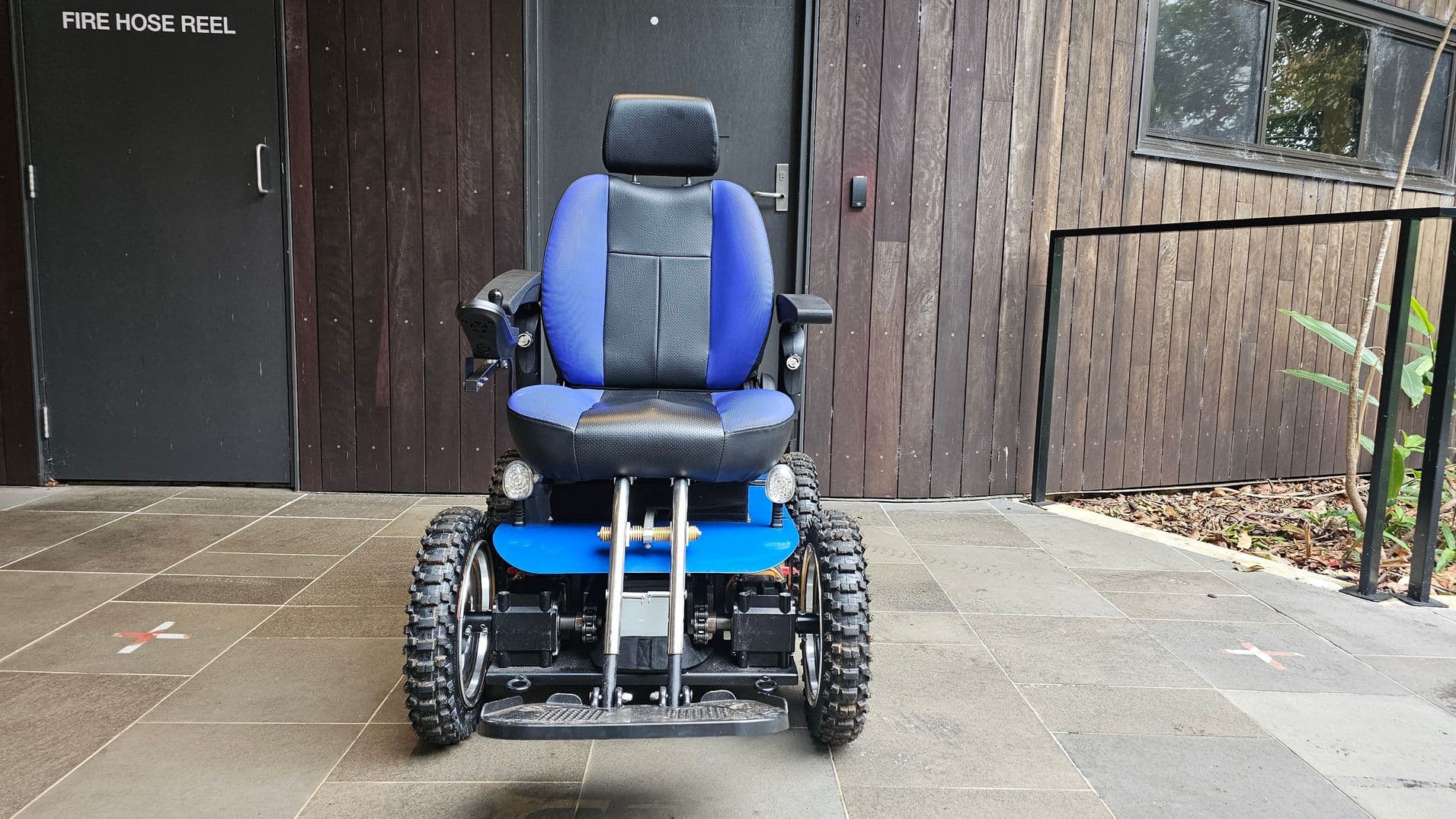 A blue and black all-terrain wheelchair with large, rugged tyres is positioned on a tiled floor. To the left, a black door with "FIRE HOSE REEL" written on it. The background features a wooden wall with a window on the right, partially reflecting greenery. A black metal railing runs along the right side, with some plants visible near the base.