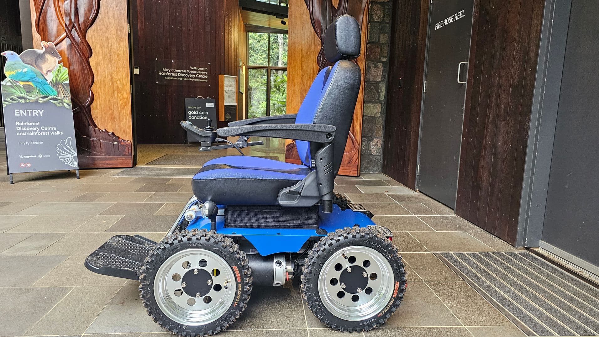 A blue and black all-terrain wheelchair with large, rugged tyres is positioned on a tiled floor. To the left, there's a wooden carved pillar and a sign for the Rainforest Discovery Centre, featuring images of a bird and a mammal. In the background, a glass door reveals greenery outside. On the right, a dark wooden wall with a door labelled "FIRE HOSE REEL" is visible.